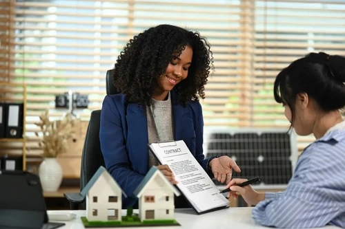 A smiling real estate agent shows a contract to a client signing paperwork at a desk with model houses on display.