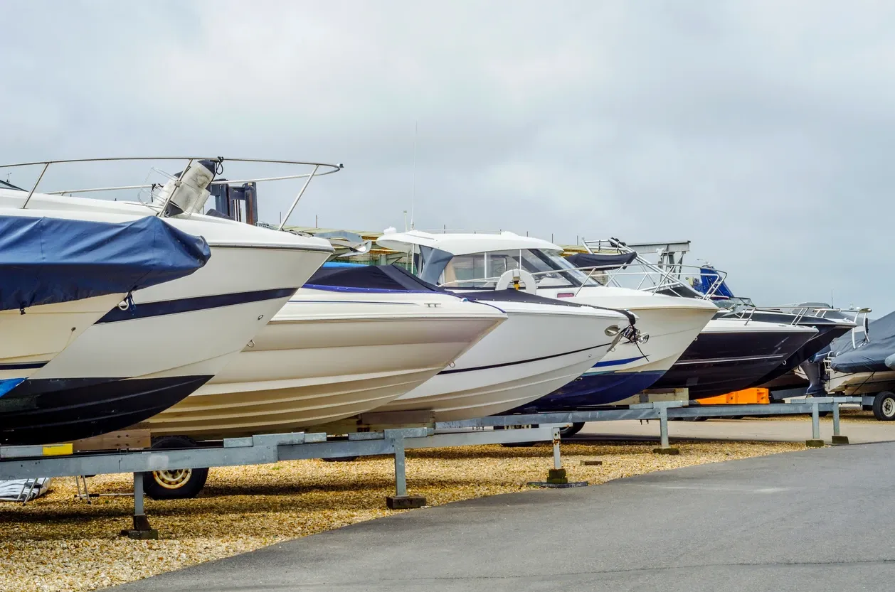 Speed boats stored in a row on brown grass just off pavement.