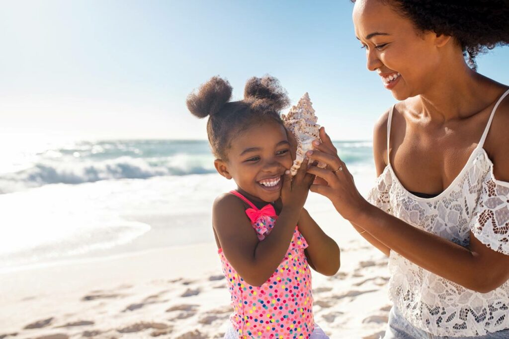 Young mother holding seashell near happy daughter’s ear at the beach while she listens to the sound of the ocean