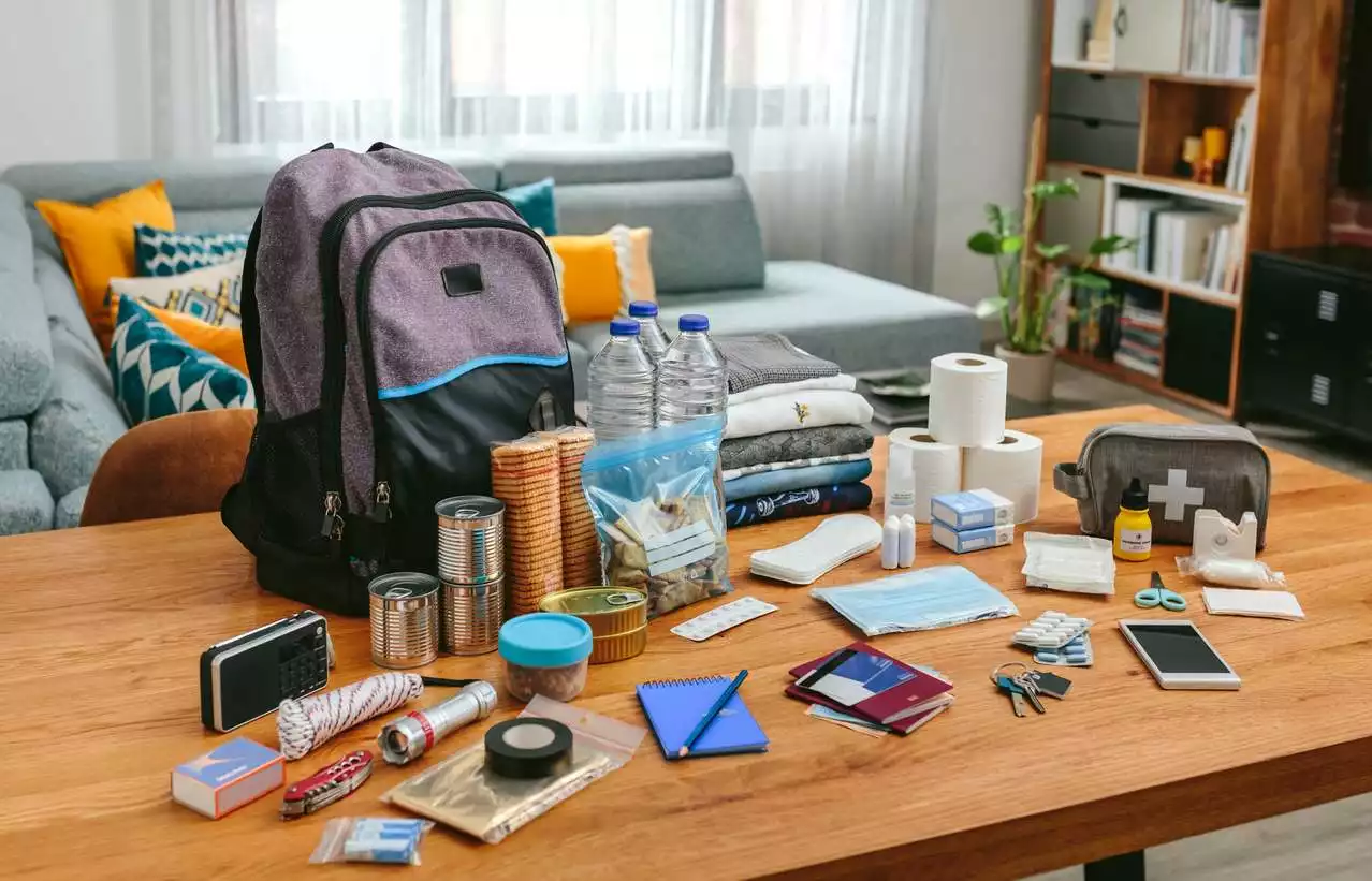 A backpack and emergency supplies, including a first aid kit, water bottles, food, and more, sitting on a table.