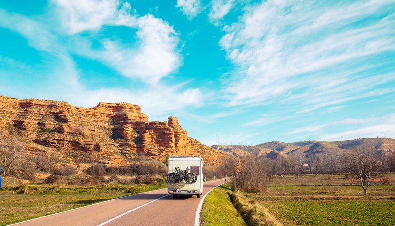 An RV driving past scenic rock formations and mountains in the midday sun.