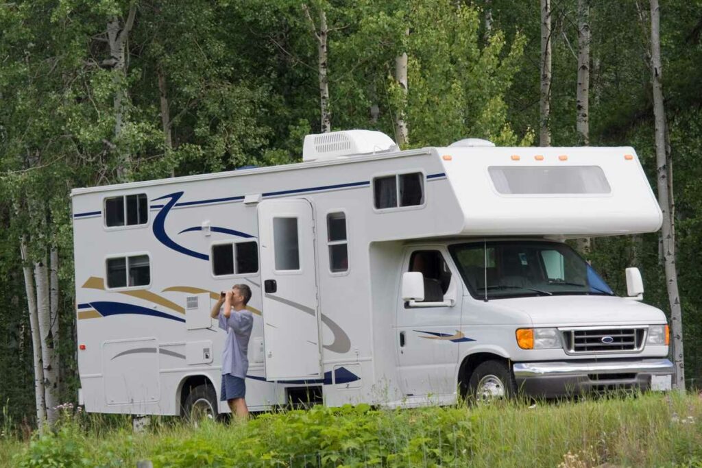 A man looks out into the woods with his large motor home. 