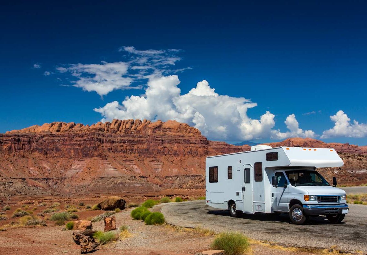 A white RV parked on a scenic desert road with red rock formations and a bright blue sky with clouds in the background.