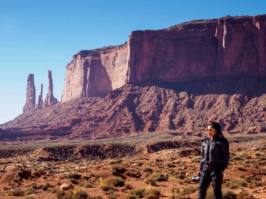 A woman in sunglasses stands in Monument Valley, with towering red rock formations and a clear blue sky. 