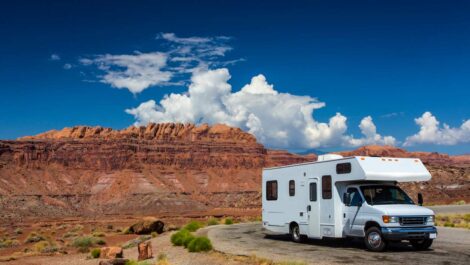 A white RV parked on a scenic desert road with red rock formations and a bright blue sky with clouds in the background.