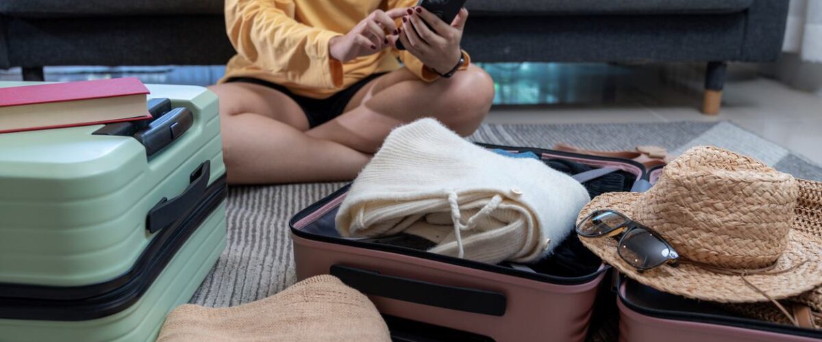 A young woman sits on the floor packing a suitcase while using her phone to plan her first road trip.