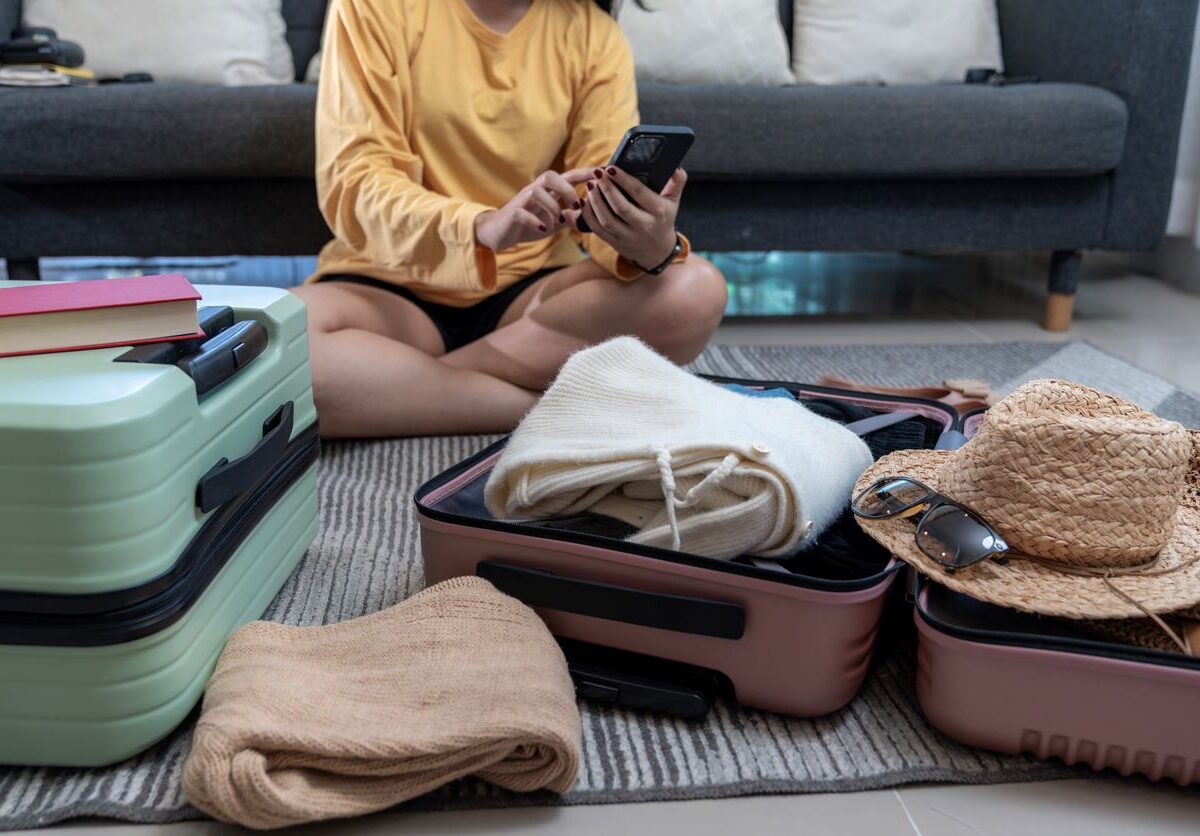 A young woman sits on the floor packing a suitcase while using her phone to plan her first road trip.