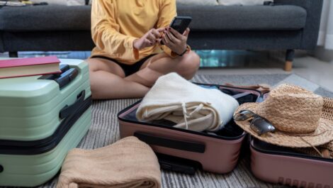 A young woman sits on the floor packing a suitcase while using her phone to plan her first road trip.