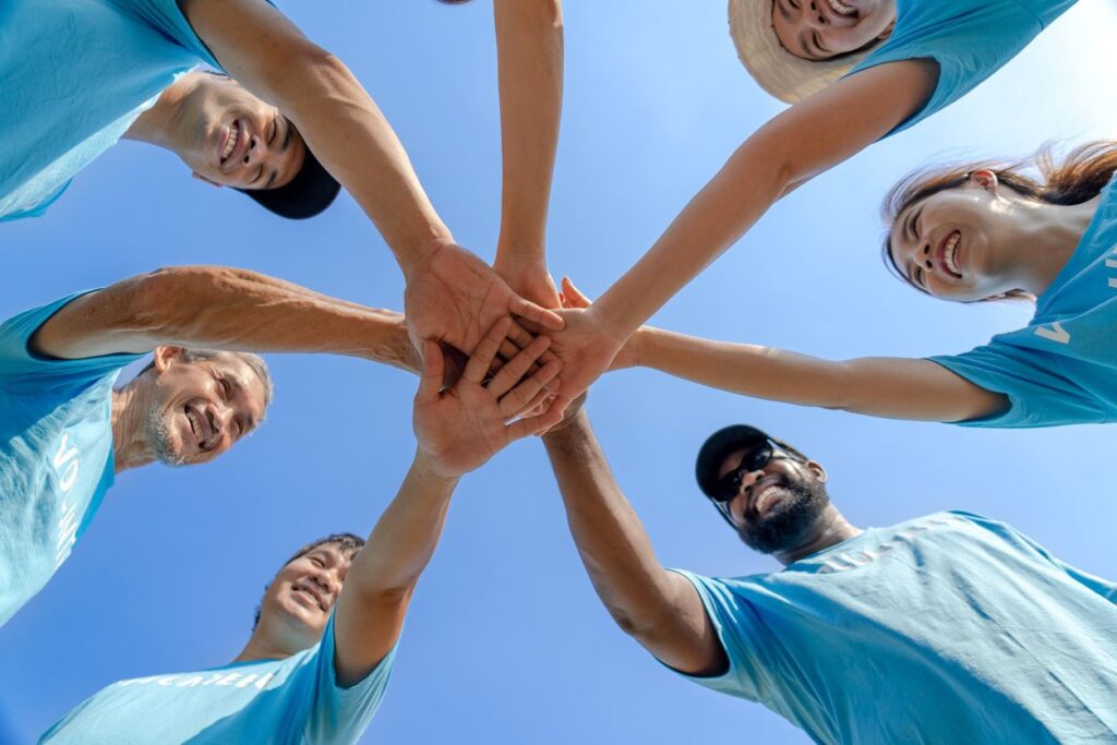 A group of volunteers in blue shirts standing in a circle with hands stacked together under a clear blue sky. 