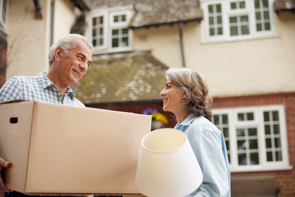 Smiling older couple carrying a box and a lamp while moving into a new home. 