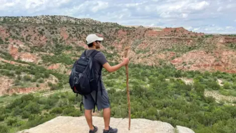 A hiker with a backpack and walking stick stands on a rocky ledge overlooking Palo Duro Canyon State Park.