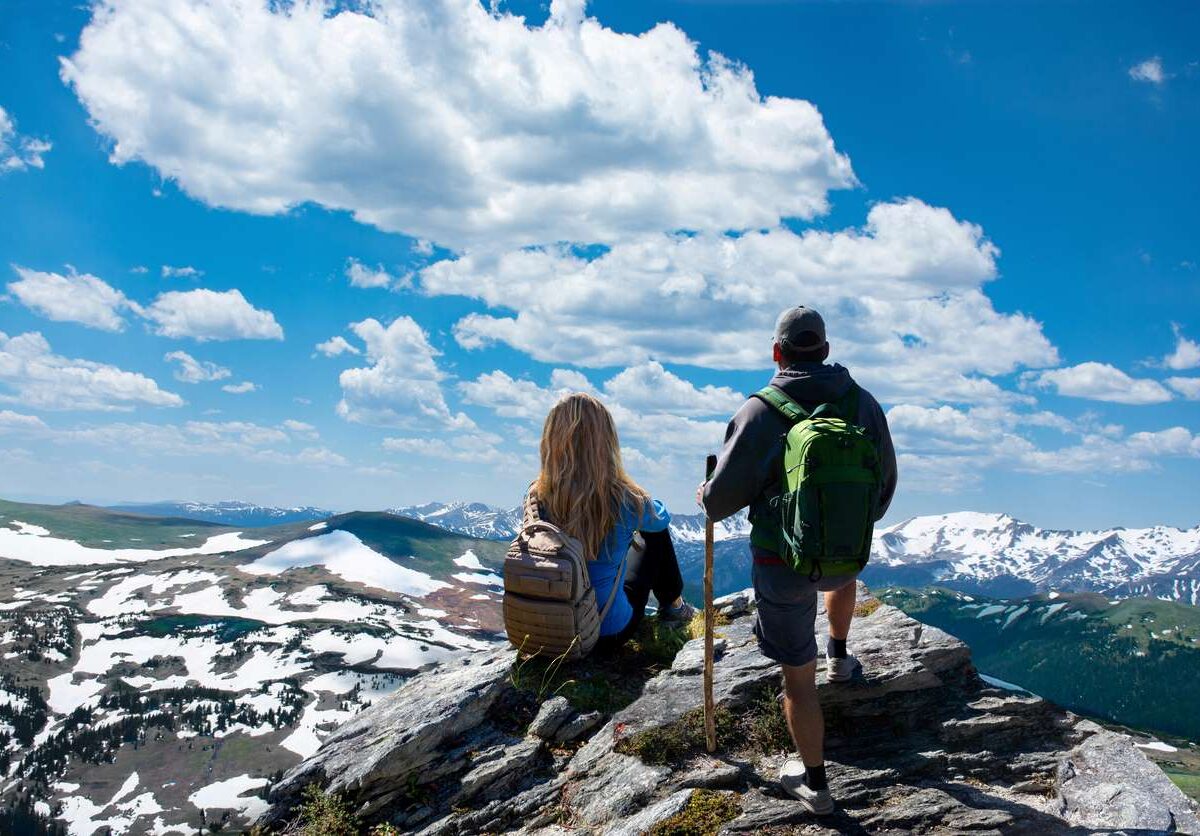 Two hikers with backpacks admire the snow-capped mountains and blue sky in the Rocky Mountains.