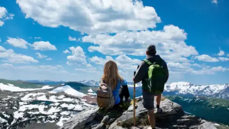Two hikers with backpacks admire the snow-capped mountains and blue sky in the Rocky Mountains.