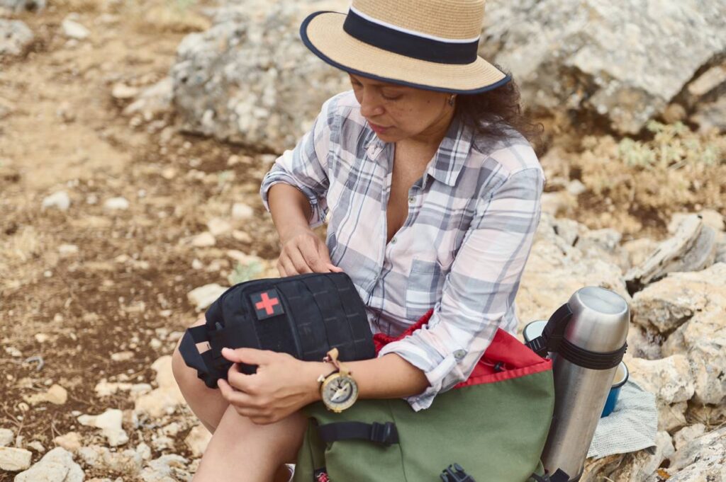 A woman in a hat and plaid shirt sitting on rocky terrain, opening a first aid kit with a backpack and thermos. 