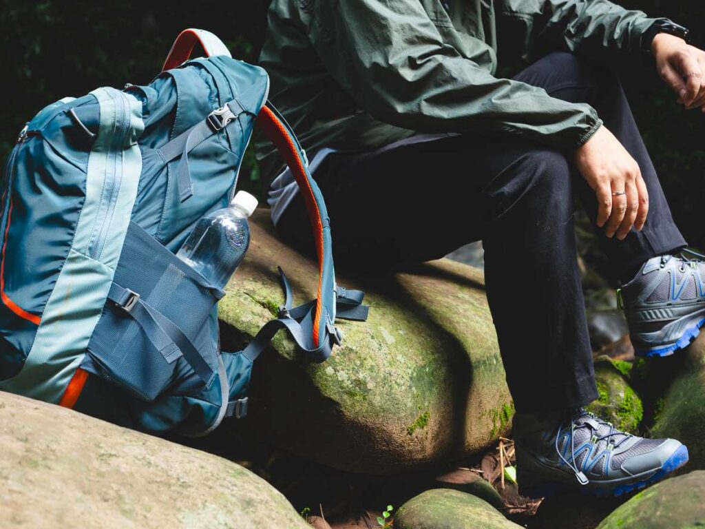 A hiker rests on rocks with a backpack, water bottle, and shoes. 