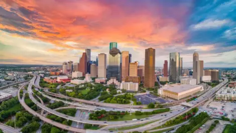 A sunset behind downtown Houston, Texas, with skyscrapers, highways, and lush green spaces.