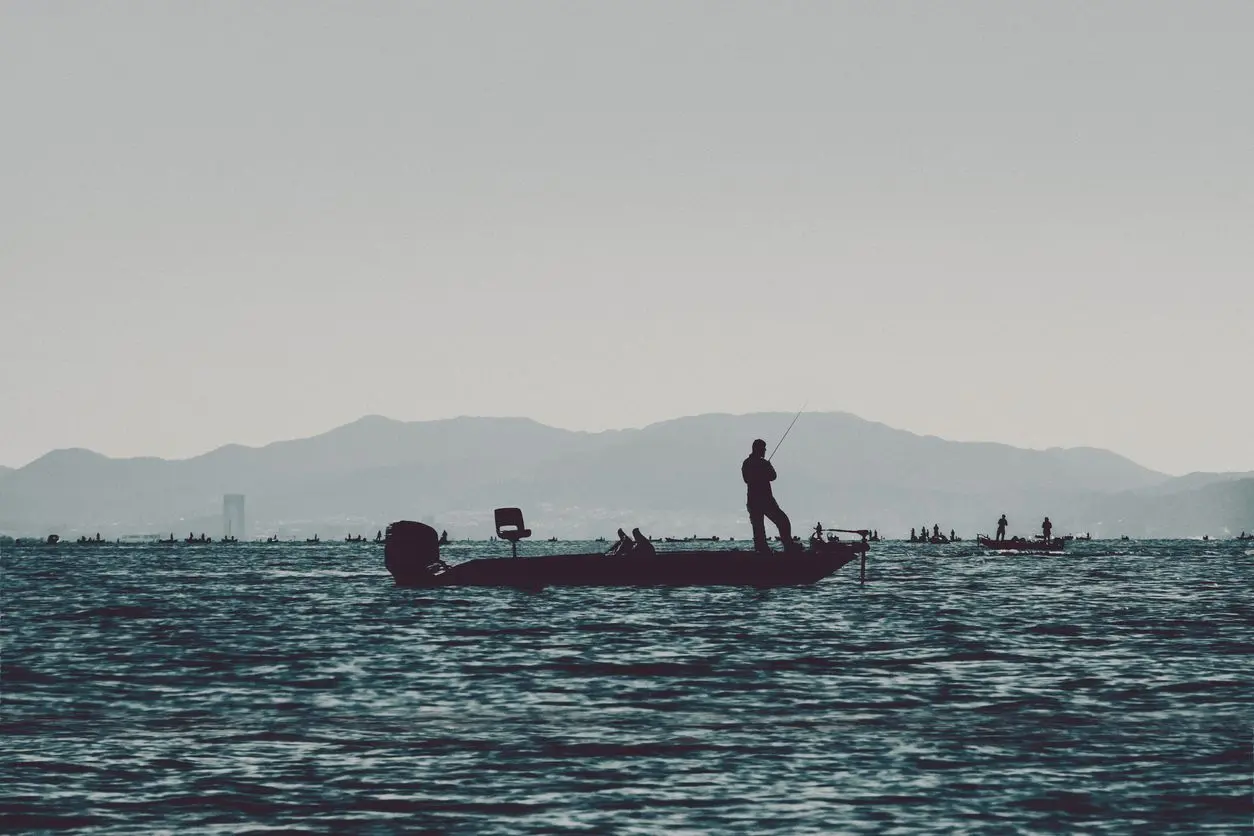  A silhouette of a man fishing on a bass boat on a lake. 