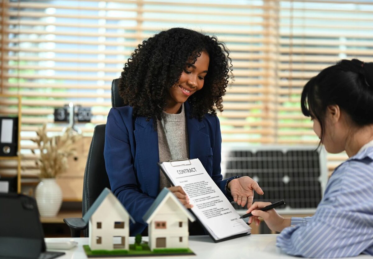 A smiling real estate agent shows a contract to a client signing paperwork at a desk with model houses on display.