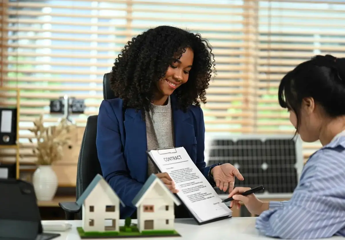 A smiling real estate agent shows a contract to a client signing paperwork at a desk with model houses on display.