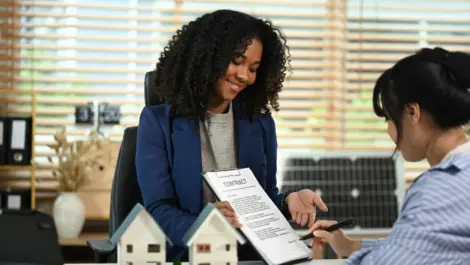 A smiling real estate agent shows a contract to a client signing paperwork at a desk with model houses on display.