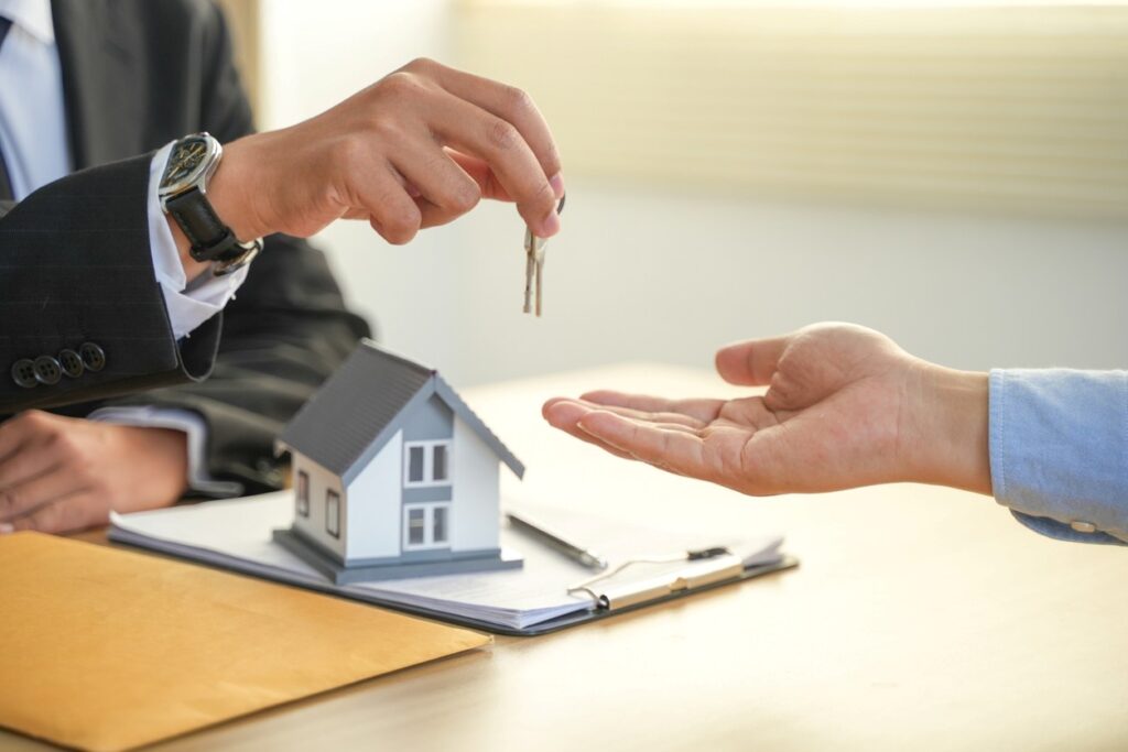 A real estate agent hands over house keys to a buyer next to a model home and paperwork on a desk.