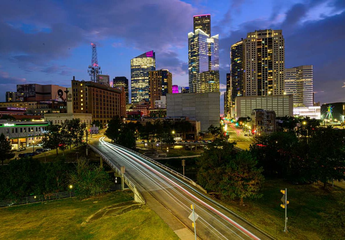 The Downtown Houston skyline at sunset with glowing skyscrapers and a vibrant purple and blue sky.