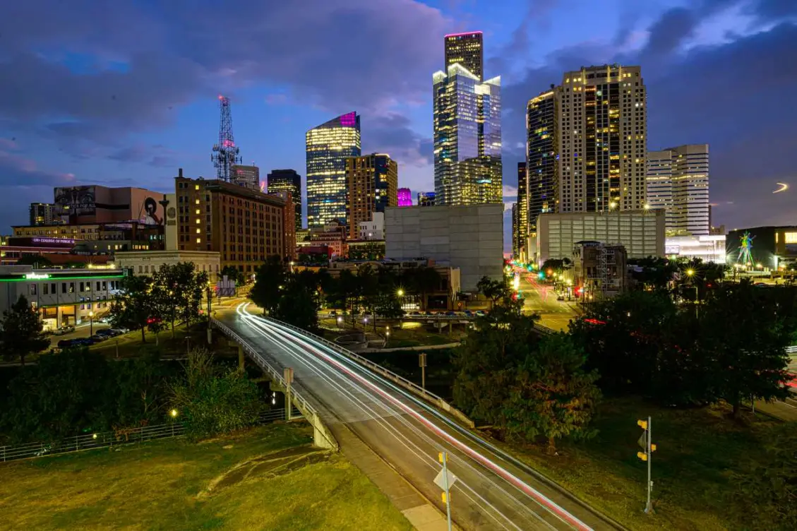 The Downtown Houston skyline at sunset with glowing skyscrapers and a vibrant purple and blue sky.
