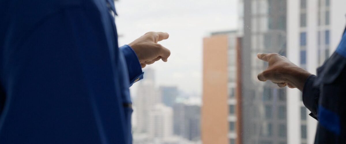 Two workers in blue uniforms point out a high-rise window toward buildings in a city skyline on a cloudy day.