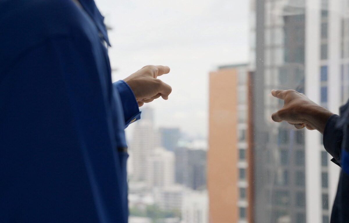 Two workers in blue uniforms point out a high-rise window toward buildings in a city skyline on a cloudy day.