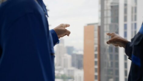 Two workers in blue uniforms point out a high-rise window toward buildings in a city skyline on a cloudy day.