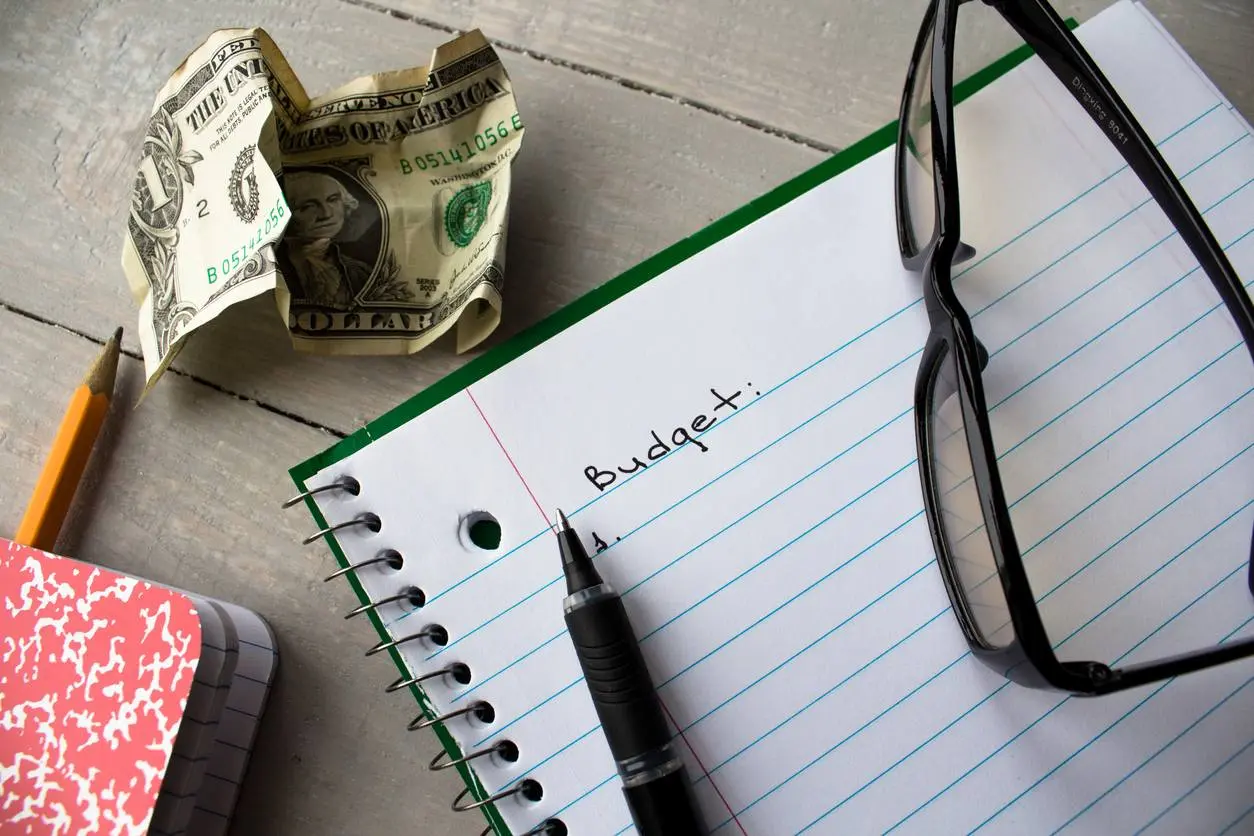 A budget planning setup with a notebook, pen, glasses, pencil, crumpled dollar bill, and journal on a wooden table. 
