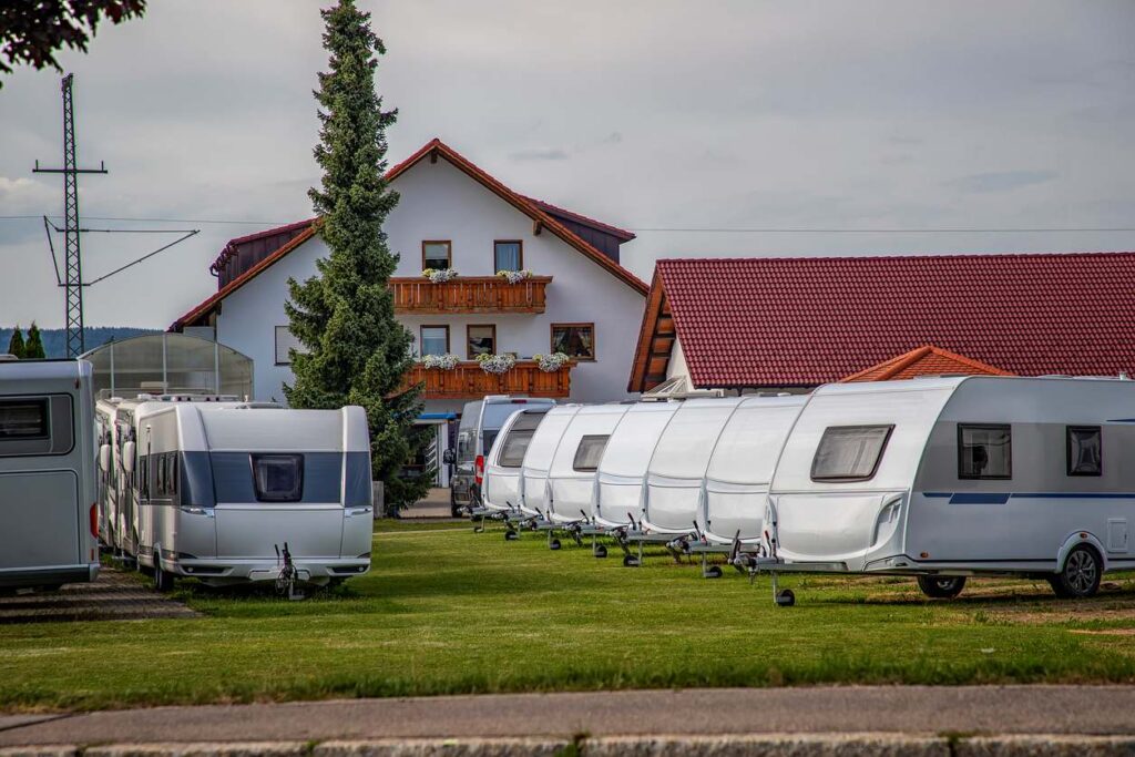 A row of parked travel trailers on a grassy lot near a house with red roofs and balconies on a cloudy day. 