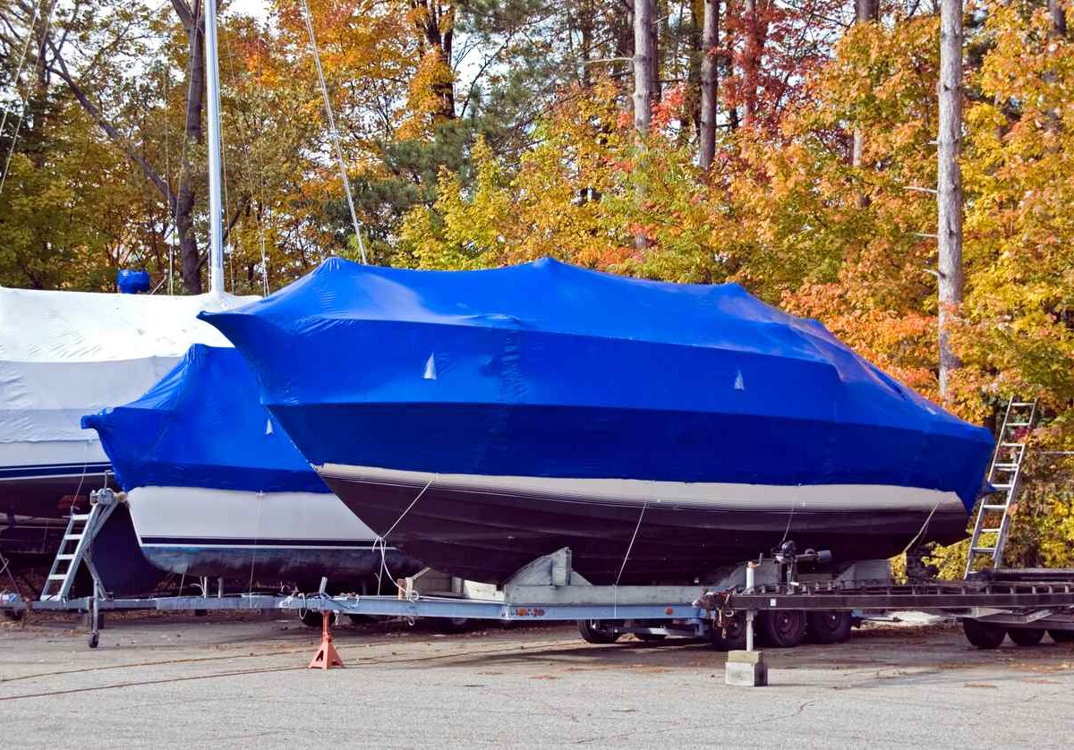 A tarp-covered boat sitting in an outdoor storage parking lot with colorful fall trees behind it.