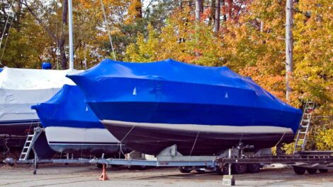 A tarp-covered boat sitting in an outdoor storage parking lot with colorful fall trees behind it.