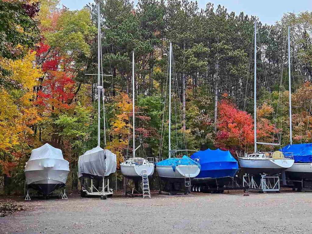 Boats wrapped in blue and white protective covers are parked on trailers in a parking lot during the autumn season. 