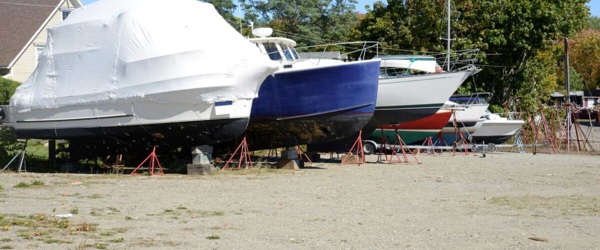 A row of boats on cement blocks parked on a dirt parking area surrounded by trees on a sunny day.