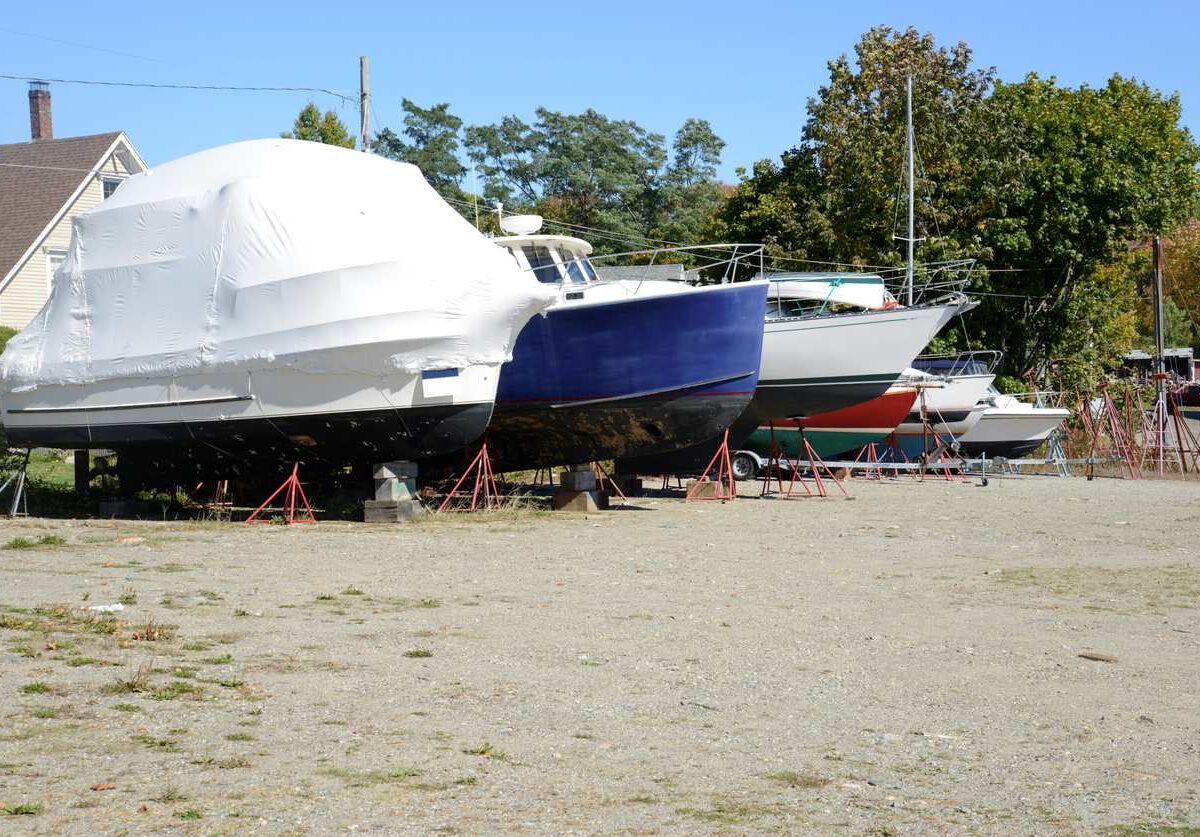 A row of boats on cement blocks parked on a dirt parking area surrounded by trees on a sunny day.