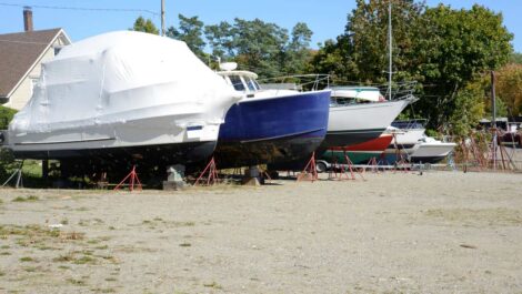 A row of boats on cement blocks parked on a dirt parking area surrounded by trees on a sunny day.
