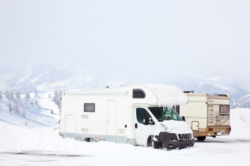 Snow-covered RVs parked in a snowy mountain area, with icicles hanging from the roof and heavy snow piled around. 