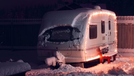 A snow-covered RV with holiday lights glowing at night, showing heavy winter buildup on the roof and windows.